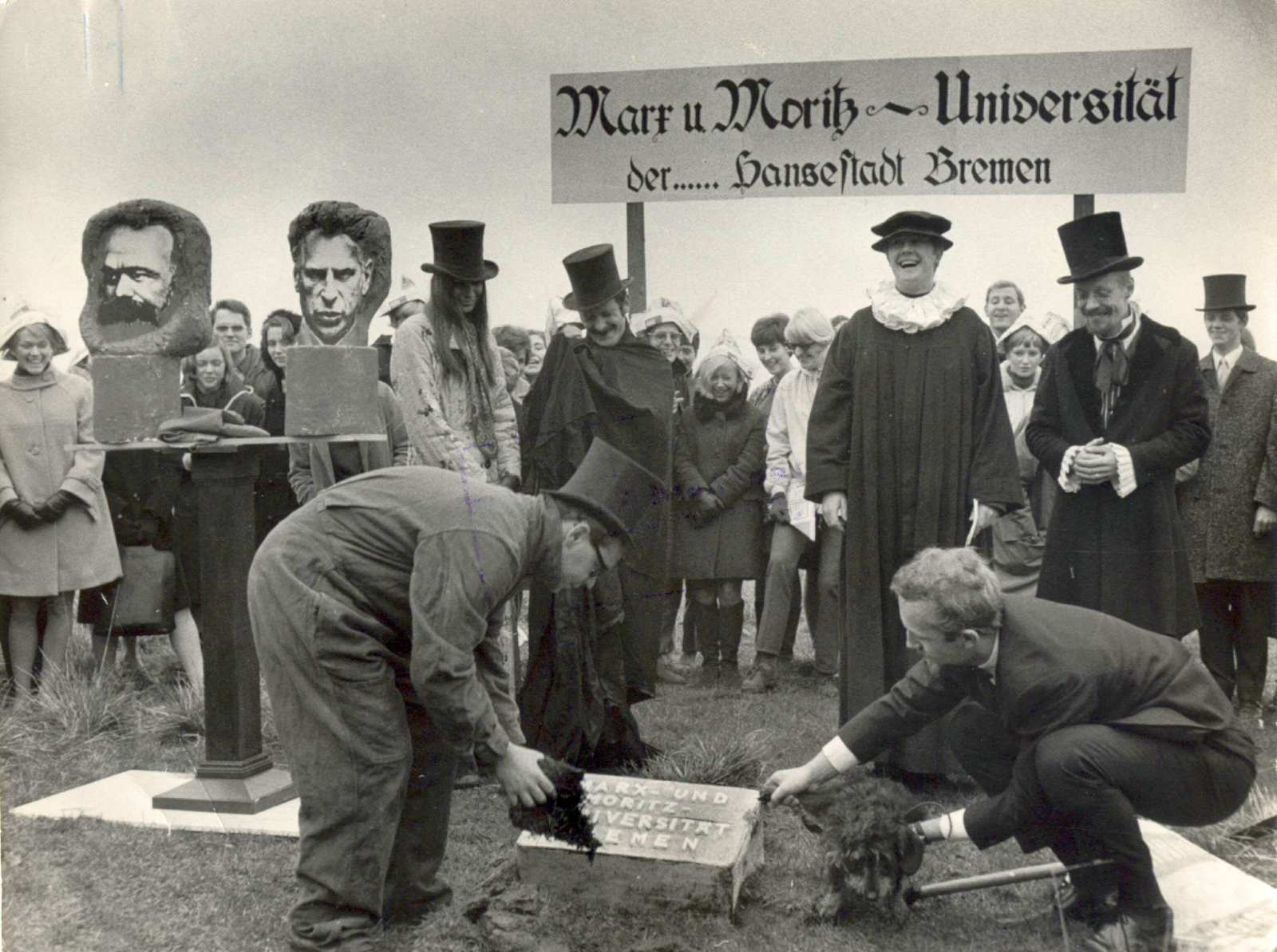 Im Vordergrund setzen zwei Männer den Grundstein für die Universität in die Wiese ein. Ein Dackel wird von einem der Männer genötigt, sein Bein an ebendiesem Grundstein zu heben. Links stehen auf einem Sockel die Büsten von Karl Marx und Moritz Thape. Lin