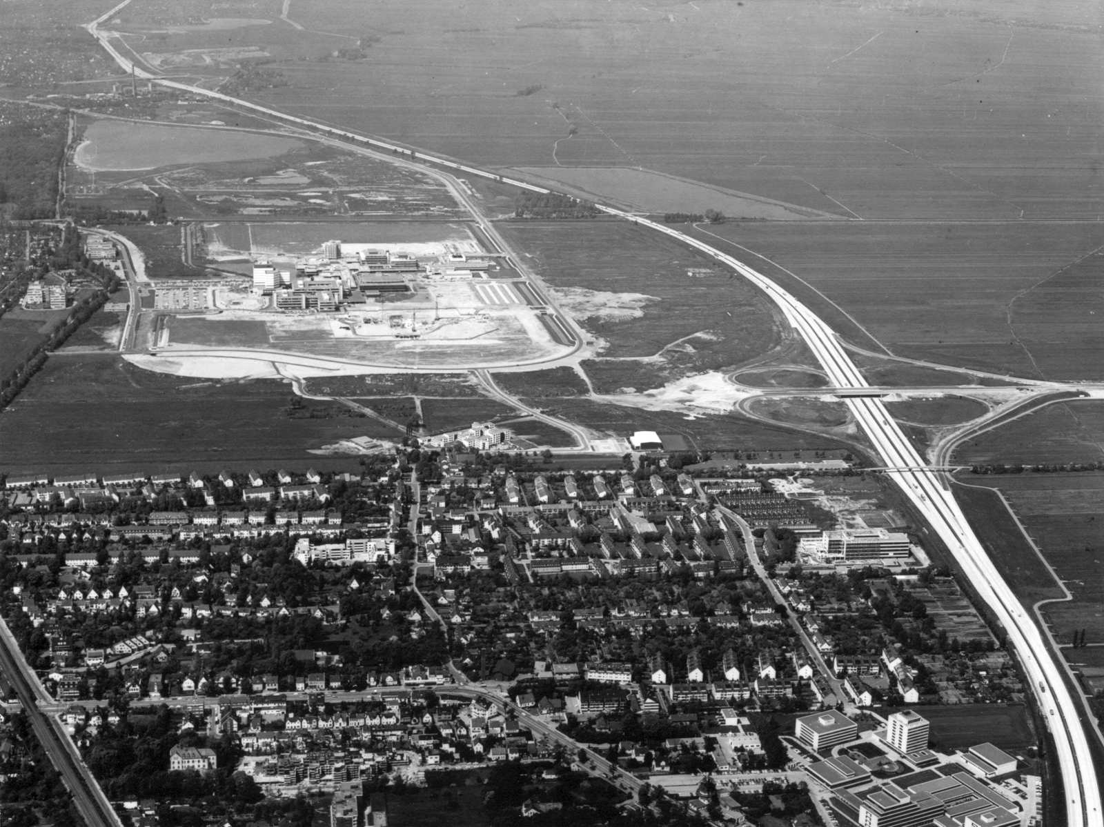 Eine Luftbildaufnahme zeigt den Campus der Universität im Jahr 1975. Im Westen begrenzt der Uni-See das Areal, im Osten zieht eine Reihe Wohnhäuser von Horn-Lehe eine klare Grenze. Die Autobahn begrenzt das Areal nach Norden. Auf der gegenüberliegenden Se