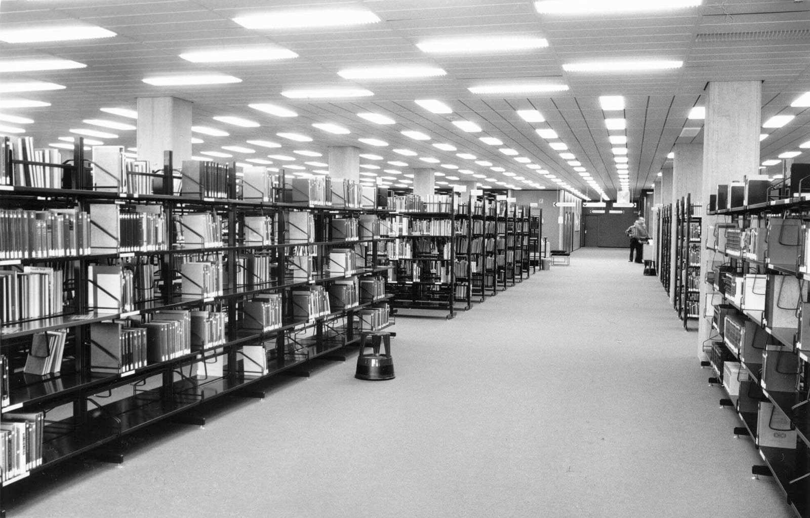 Ein hell erleuchteter Raum mit einem breiten Gang an dem rechts und links mit Büchern gefüllte Regale stehen und weitere Gänge abgehen. Am Ende des breiten Ganges stehen zwei Männer an einer Informationstheke.
