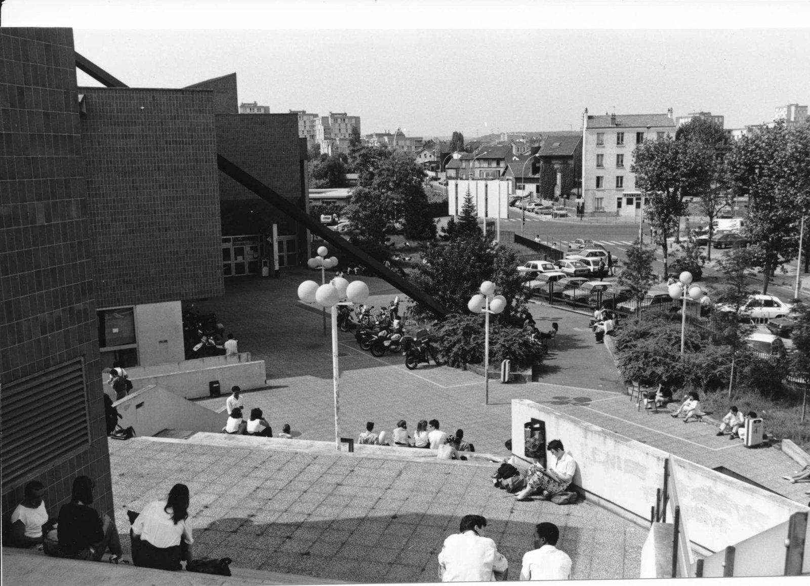 Blick entlang einiger Gebäude der französischen Universität. Auf den Treppenstufen sitzen Studierende. Im Hintergrund sind Parkplätze, Straßen und Wohnhäuser.