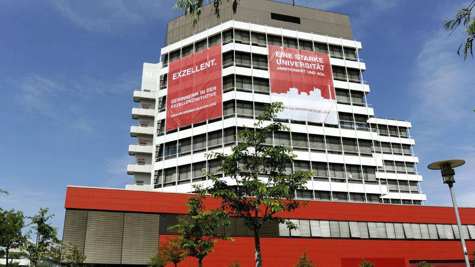 Das Gebäude MZH mit zwei großen roten Bannern. Sie tragen die Aufschrift: Exzellent. Gewinnerin der Exzellenzinitiative. Und: Eine starke Universität.