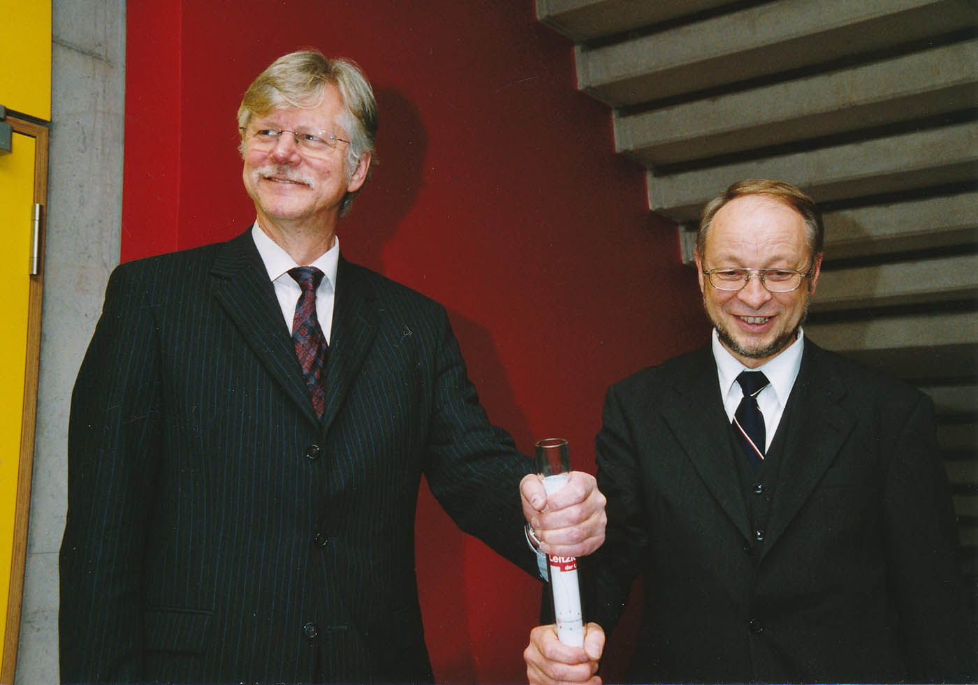 Links steht Wilfried Müller mit einer Hand am Acrylglasstab mit den Leitzielen der Uni. Rechts daneben steht Alt-Rektor Jürgen Timm auch mit einer Hand am Stab.