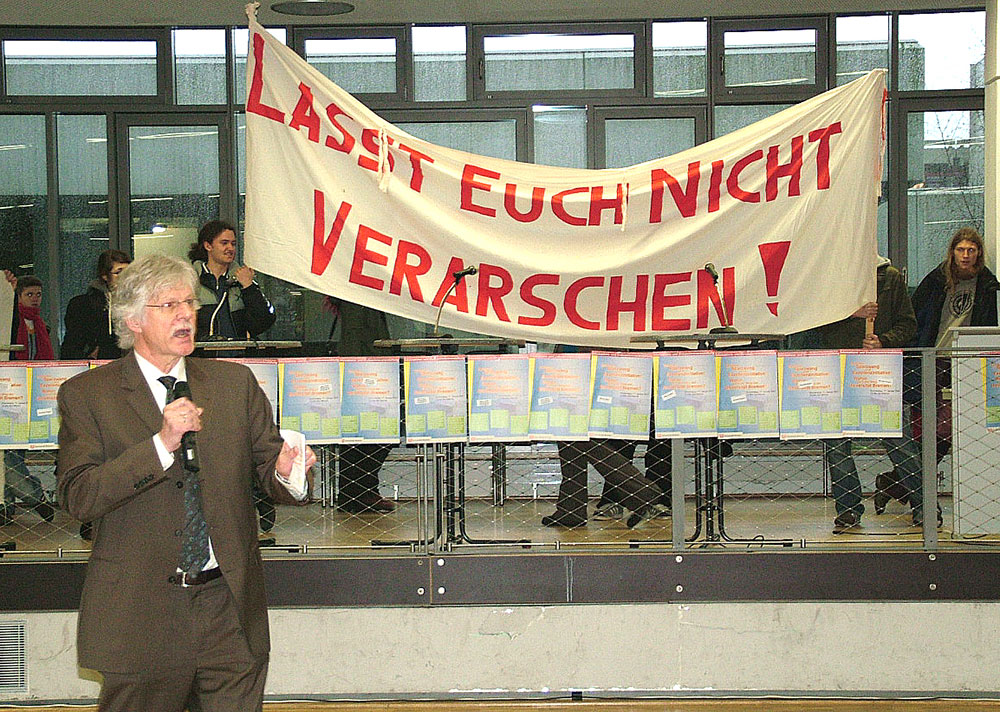 Rektor Wilfried Müller steht neben einem Spruchband mit der Aufschrift „Lasst euch nicht verarschen“ und spricht sichtlich erregt in ein Mikrofon.