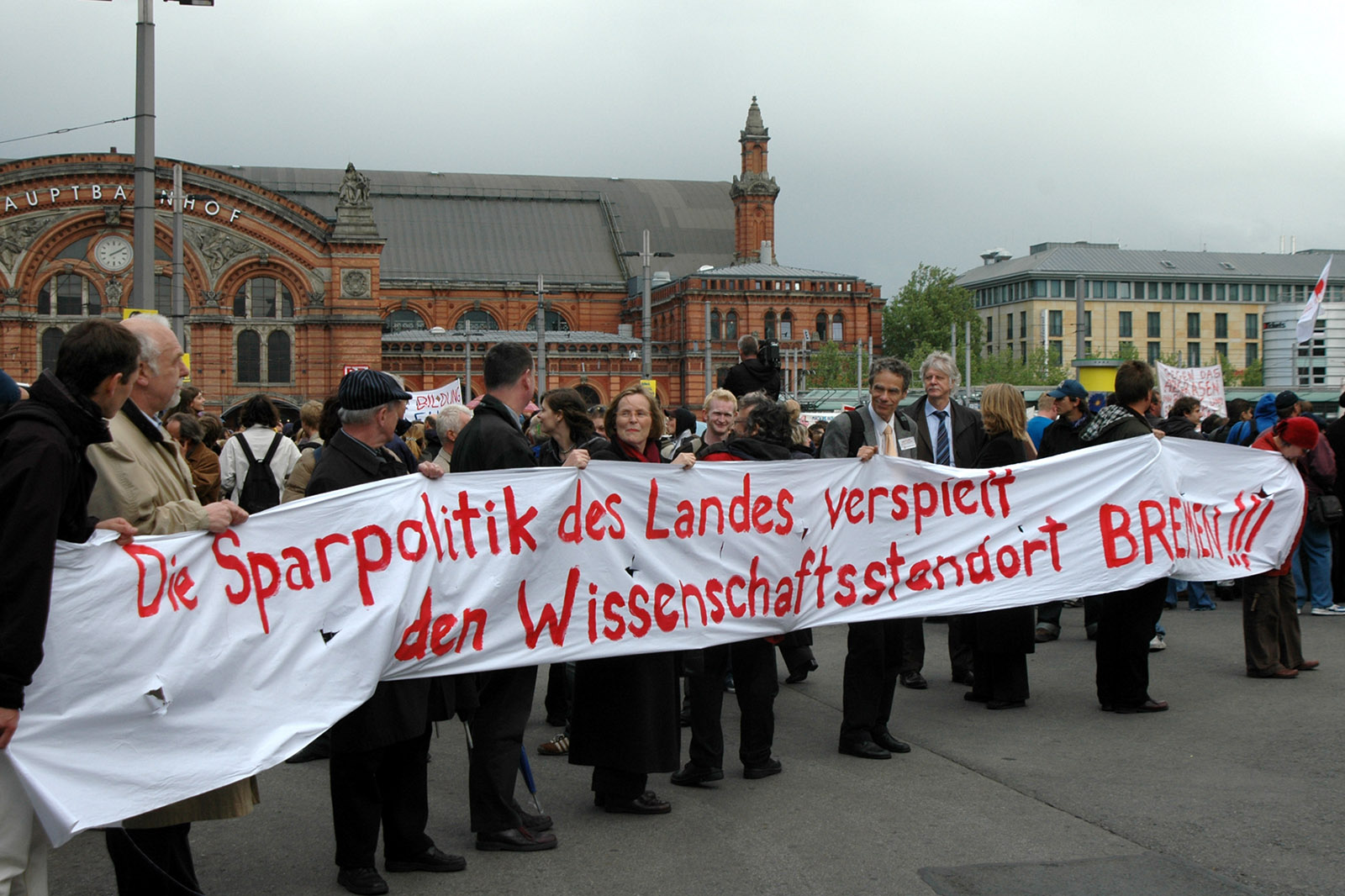 Vor dem Bremer Bahnhof steht eine Gruppe von Menschen, die ein Spruchband hochhalten mit dem Worten: „Die Sparpolitik des Landes verspielt den Wissenschaftsstandort Bremen“.