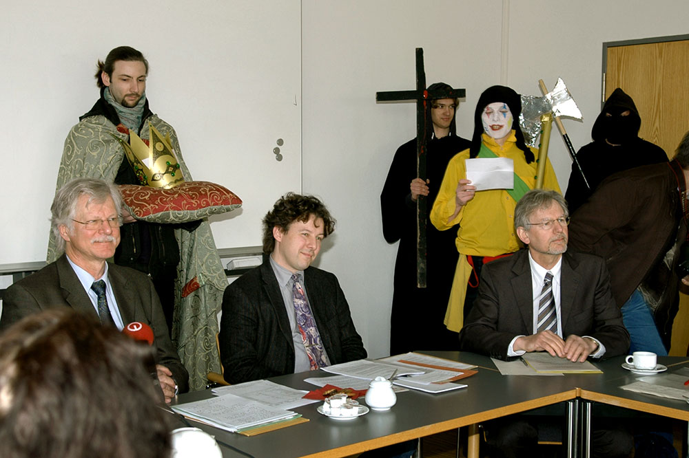 An einem Tisch sitzt Rektor Wilfried Müller mit weiteren Personen. Vor ihnen stehen Mikrofone, auf denen Logos von Medienanstalten zu sehen sind. Hinter ihnen haben sich verkleidete und geschminkte Studierende aufgestellt.