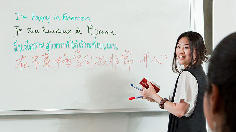 Eine Frau steht an einem Whiteboard, auf dem Wörter in verschiedenen Sprachen stehen.